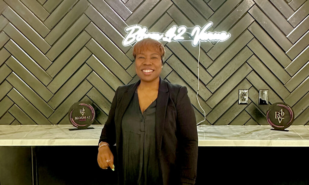 Atiya Hobson, a Black woman, stands in front of a neon sign that says Bloom 42
